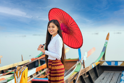 A young Burmese woman holding a red umbrella with the gondola and looking camera. U-bein bridge, Mandalay, Myanmar.