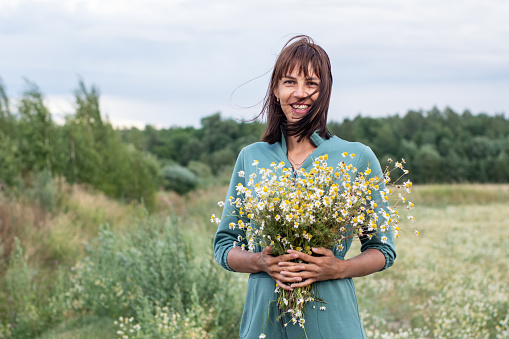 Portrait of a young woman of European appearance in a summer field. A woman with a bouquet of daisies in a chamomile field. A bouquet of wild flowers in your hands