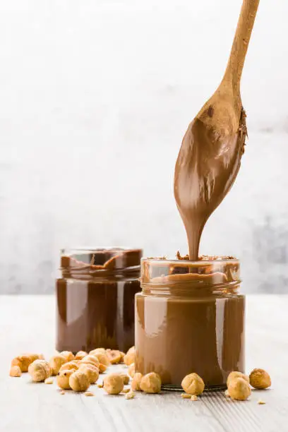 Chocolate cream in glass jar with hazelnuts and dripping spoon, on wooden table