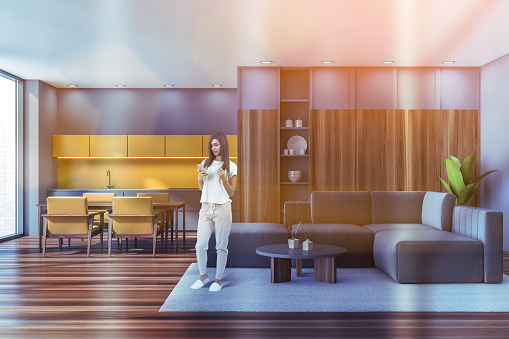 Attractive young woman with phone in wooden modern living room with kitchen, lens flare. Couch, coffee table and panoramic windows.