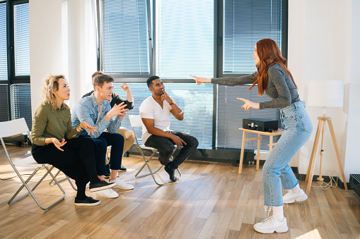 Side view of cheerful redhead woman playing charades with friends showing pantomime by window in office. Group of activity diverse multi-ethnic colleagues playing in active games during team building.
