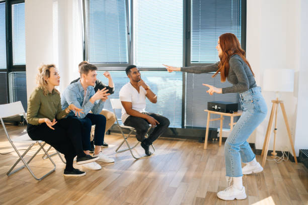 Side view of cheerful redhead woman playing charades with friends showing pantomime by window in business office. Side view of cheerful redhead woman playing charades with friends showing pantomime by window in office. Group of activity diverse multi-ethnic colleagues playing in active games during team building. pantomime stock pictures, royalty-free photos & images