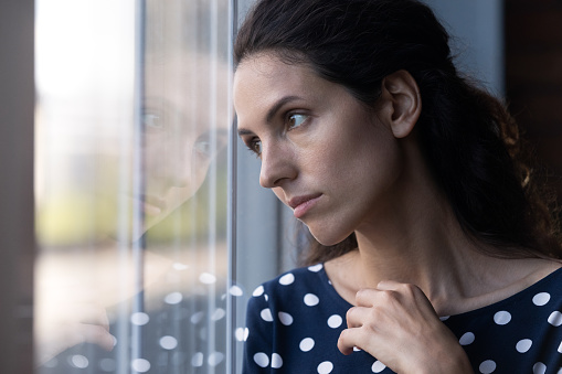 Sad thoughtful Hispanic woman looking out window in deep thought, thinking over difficult heartbreak, feeling bored, melancholy, apathy, suffering from negative mood, depression, going through trauma