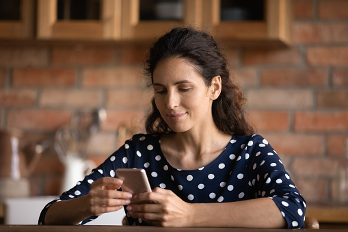 Focused Latin woman reading text on cellphone screen in kitchen at home, using online app, virtual service, ordering food delivery, making call, quick payment, chatting on social media