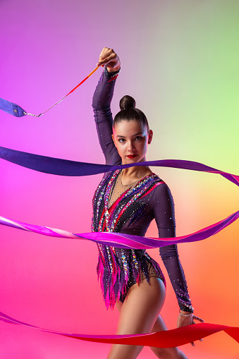 Half length portrait of one young rhythmic gymnast posing with ribbon in her hand isolated over bright multicolored neon background. Concept of sport life, motion, flexibility.