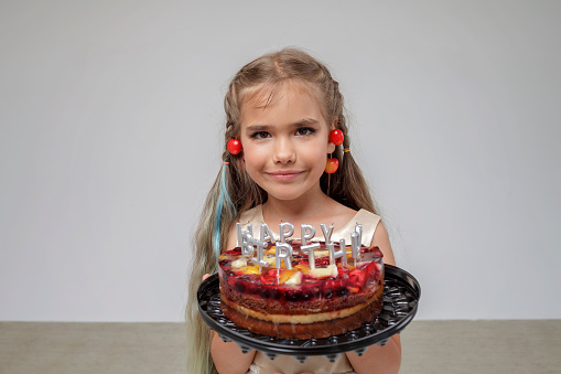 Preteen girl with cherry on the ears holding birthday cake with candles and blowing them out over white pastel background, birthday celebration party, close-up portrait, studio shot