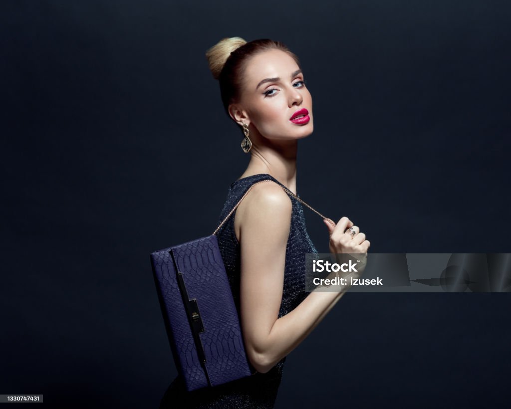 Portrait of elegant woman holding hand bag Glamour portrait of elegant, beautiful woman wearing black evening gown and holding purple purse. Studio shot against black background. Evening Bag Stock Photo