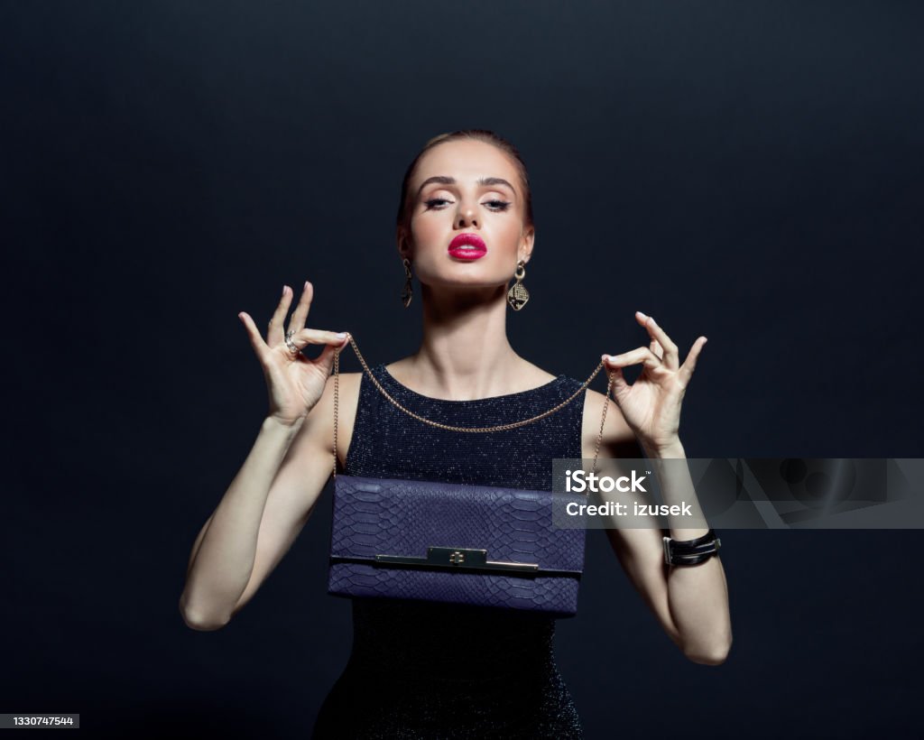 Portrait of elegant woman holding hand bag Glamour portrait of elegant, beautiful woman wearing black evening gown and holding purple purse. Studio shot against black background. 20-29 Years Stock Photo