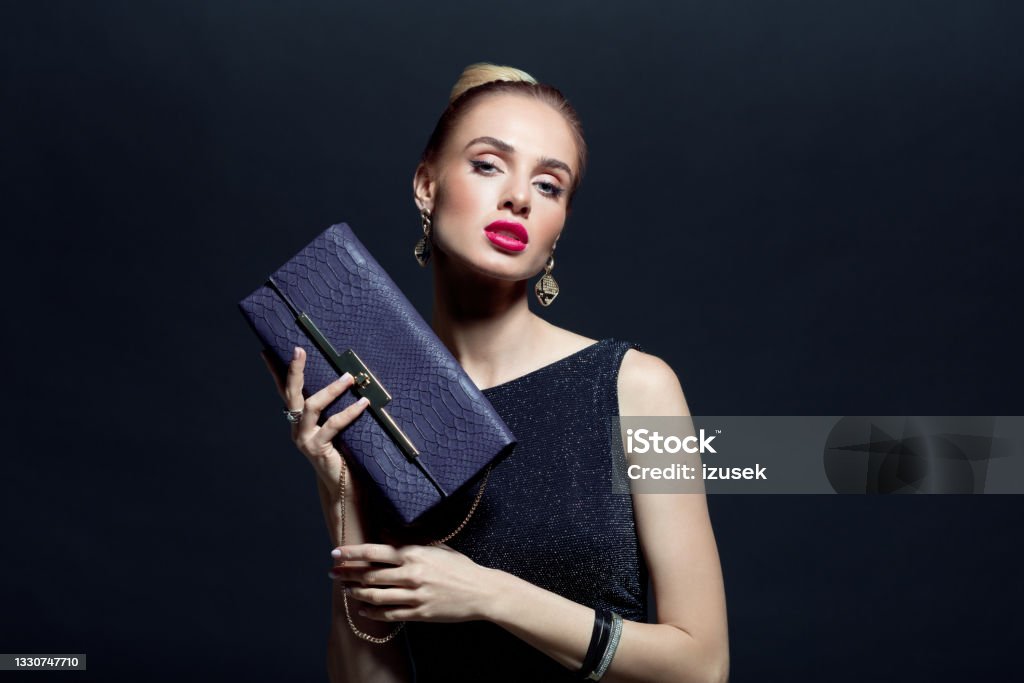 Portrait of elegant woman holding hand bag Glamour portrait of elegant, beautiful woman wearing black evening gown and holding purple purse. Studio shot against black background. Purse Stock Photo