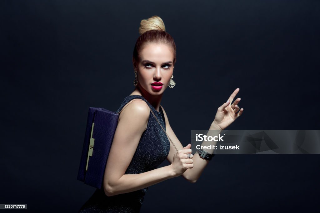 Portrait of elegant woman holding hand bag Glamour portrait of elegant, beautiful woman wearing black evening gown and holding purple purse. Studio shot against black background. Luxury Stock Photo
