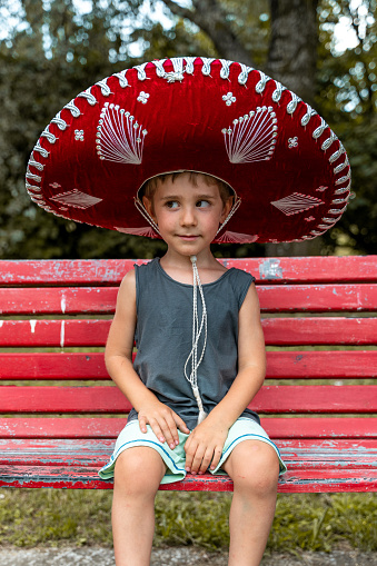 little cute boy wearing a sombrero is sitting on a red bench outdoors