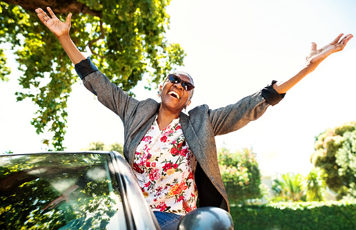 Happy senior woman enjoying car ride, getting out of window car with raised hands and laughing