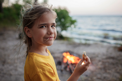 Cute girl roasting marshmallow to make smores over fire flame during camping, traditional American food, active recreation and family travel
