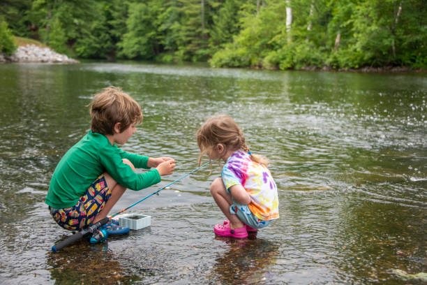 A young boy prepares his fishing rod with bait as his little sister watches intently. Preparing to fish where Red Bridge Dam empties Lake Rescue into Lake Pauline in Vermont.