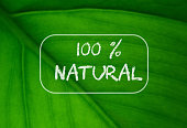 100% natural written against with green leaf blurred background. Organic, bio and eco concept for stickers, banners, cards, advertisement. Copy space.