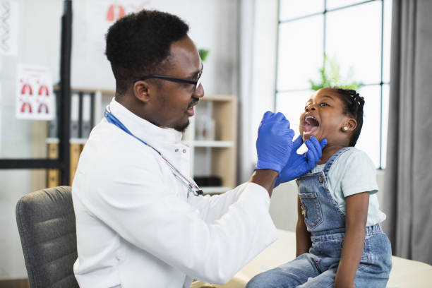 enfant afro-américaine au cabinet du médecin, beau médecin noir qui examine sa gorge - doctor patient male tongue depressor photos et images de collection