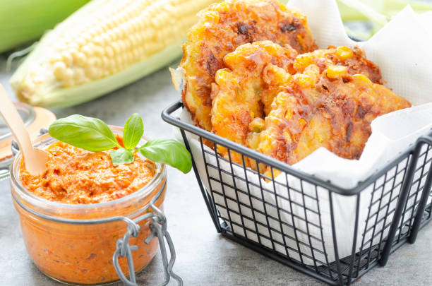 Corn fritters with red bell pepper pesto Homemade fried corn fritters made of eggs ,cheddar, milk, corn kernels,flour, seasoning and cilantro on a frying basket with red bell pepper pesto and fresh corn cob in background for summertime basket healthy eating vegetarian food studio shot stock pictures, royalty-free photos & images
