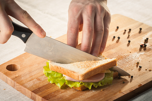 Cutting sandwich with bread, cheese, salad and ham with hands on wooden cutting board with knife.