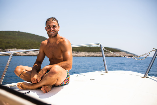 A fit young man is sitting at the bow of a powerboat, smiling at the camera