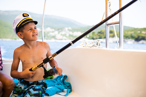 A cute boy with a ship captain's hat is enjoying fishing on a boat at the seaside