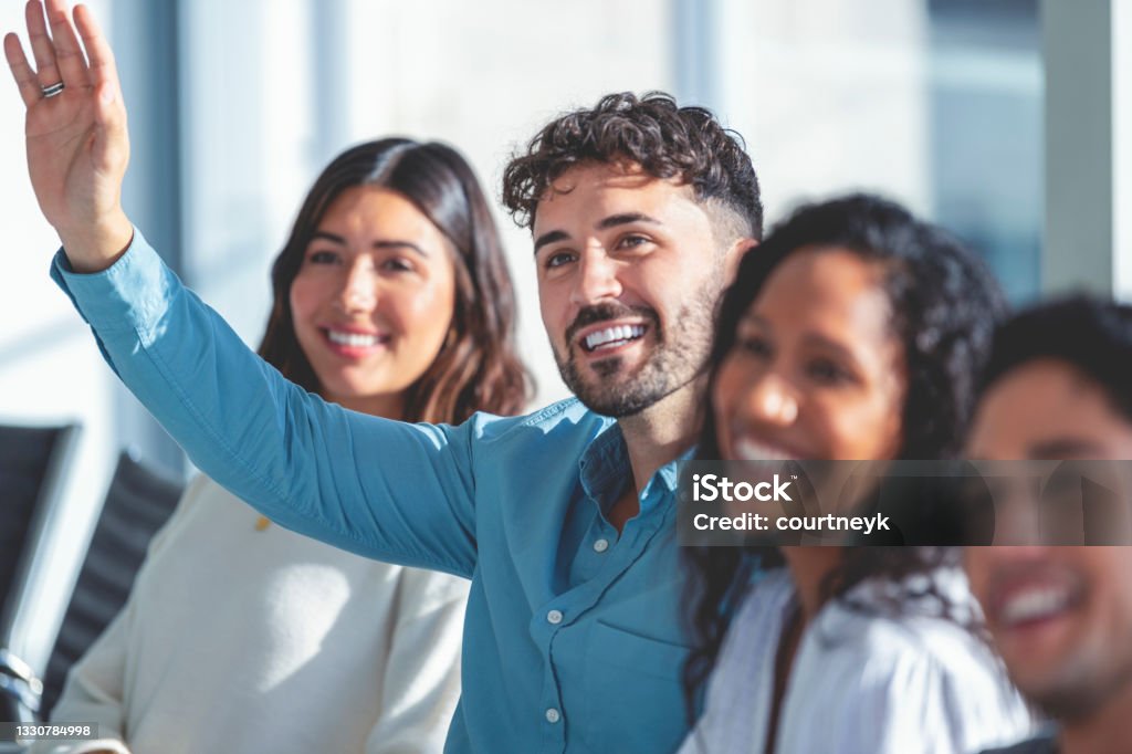 Group of people listening to a presentation. Group of people listening to a presentation. One person has their hand raised. They are at a seminar or meeting sitting at a table. They are all looking to the front of the room. Men and women dressed in casual business attire. Multi-cultural group with African, Latino and Caucasian Question Mark Stock Photo