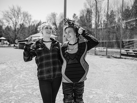 Female LGBTQ couple having a break after practising baseball at the baseball field. Young transgender woman and young  lesbian woman dressed in casual clothes with baseball gear.  Exterior of public baseball field in the park.