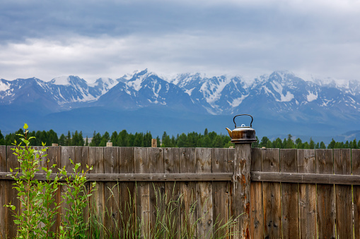 Tourist old teapot on the fence on the background of snow vertices. Summer landscape in the Altai Republic in Russia. Traveling around mountain ranges in summer.