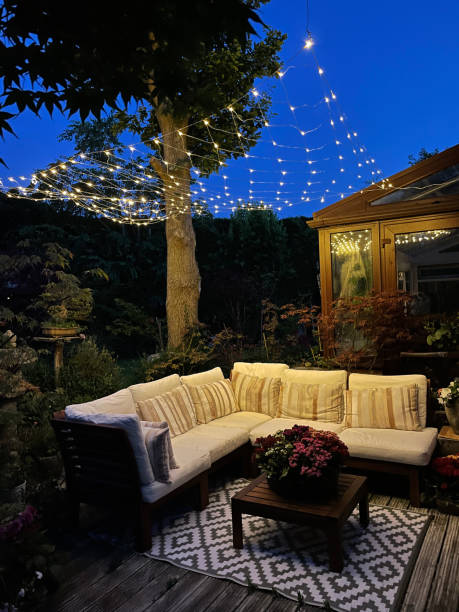 Image of outdoor lounging area at night illuminated by string fairy lights, hardwood seating with cushions, wooden table top with flowering plant centrepiece, bonsai trees, Japanese maples, landscaped oriental design garden, focus on foreground Stock photo showing ornamental Japanese-style garden with outdoor lounge area at night illuminated with fairy lights. Featuring crystal clear koi pond, whitewashed, grooved timber decking patio, Japanese bonsai maples and hardwood, cushion covered seating. garden feature stock pictures, royalty-free photos & images