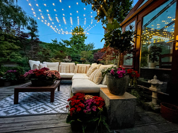 Image of outdoor lounging area at night illuminated by string fairy lights, hardwood seating with cushions, wooden table top with flowering plant centrepiece, bonsai trees, Japanese maples, landscaped oriental design garden, focus on foreground Stock photo showing ornamental Japanese-style garden with outdoor lounge area at night illuminated with fairy lights. Featuring crystal clear koi pond, whitewashed, grooved timber decking patio, Japanese bonsai maples and hardwood, cushion covered seating. patio stock pictures, royalty-free photos & images