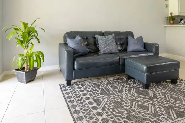 An angled view of a  large blue modem styled love seat and ottoman sofa made of genuine leather. The couch is by a large bright window and surrounded with neutral colored tile floor and wall. A geometric black and grey carpet and green plan fill the space.