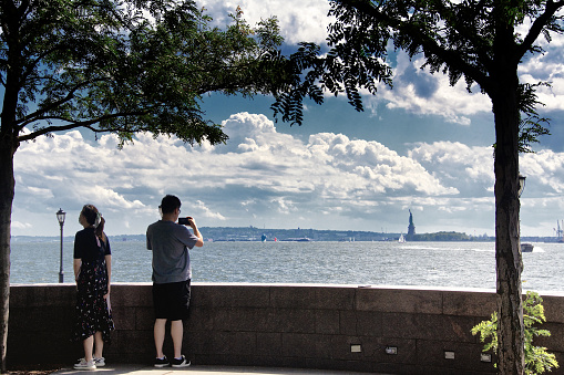 New York City, USA - July 04, 2021: At the Hudson River and Upper New York Bay, the Statue of Liberty is seen in the distance as a tourist photographs the scene  from Lower Manhattan.