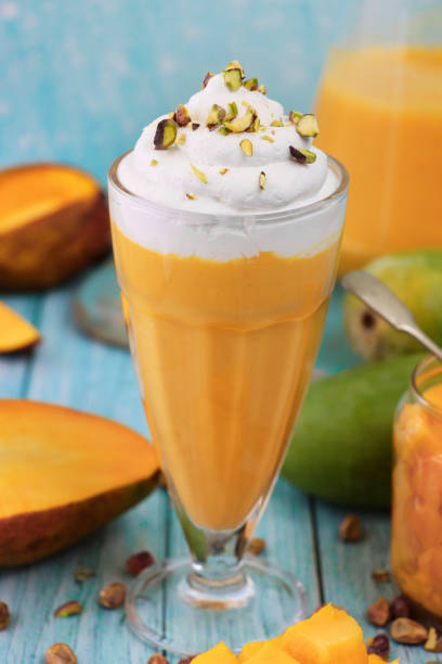 Stock photo showing mango smoothies / milkshake drinks, served in American diner-style knickerbocker glasses. These drinks have been made with pureed mango, milk and whipped cream, and topped with chopped Pistachio nuts. imagen de primer plano knickerbocker vaso de batido de mango cubierto con remolinos de crema batida y nueces de pistacho picadas además de trozos de mango en dados de jarra de vidrio, cuchara, jarra de batido de mango, sobre superficie de grano de madera