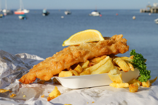 Stock photo showing close-up view of takeaway fish and chips on cardboard tray wrapped in paper with mushy pea fritter, slice of lemon and garnished with parsley. This is a fish supper being eaten at the seaside with a background of a pier and boats floating on the sea.