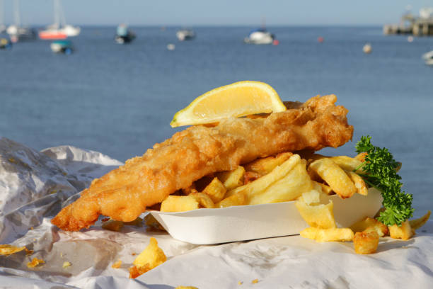 close-up image of cardboard takeaway tray of fish and chips with mushy pea fritter, slice of lemon, parsley, wrapped in paper, unhealthy food takeout, greasy battered cod and deep fried chips ready to eat at the seaside, boats floating on sea - swanage imagens e fotografias de stock