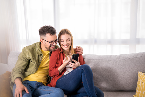 Happy young couple relaxing together on sofa while searching the internet on smart phone.