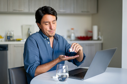 Latin American man working at home and taking pills for a headache - healthcare and medicine concepts