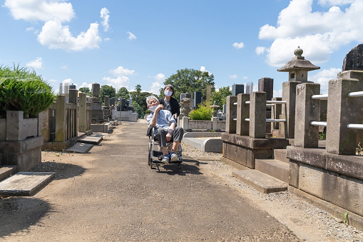 Elderly Japanese husbund and wife who is wearing a mask visited grave. The old lady push her husbund who is getting on a wheelchair from the way back to the grave. It is a very hot summer day (Ohigan:the week of the equinox) and white clouds in the blue sky.