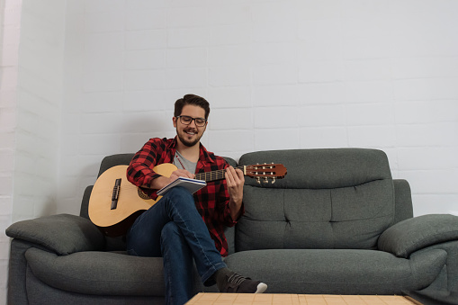 Latin hipster in glasses and red plaid shirt writing his musical composition in a notebook while holding the guitar.