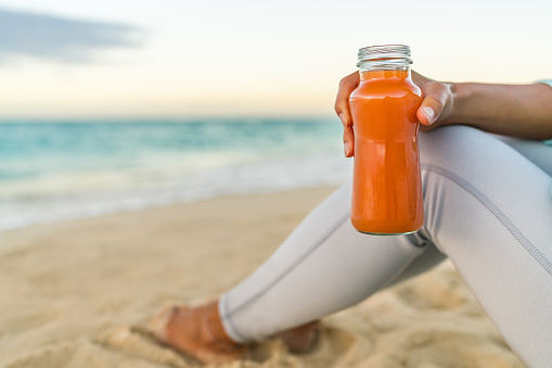 Healthy carrot vegetable juice detox cleanse woman drinking smoothie for weight loss diet at beach sunset. Closeup of fresh orange glass bottle. Juicing trend, raw, organic and cold-pressed.