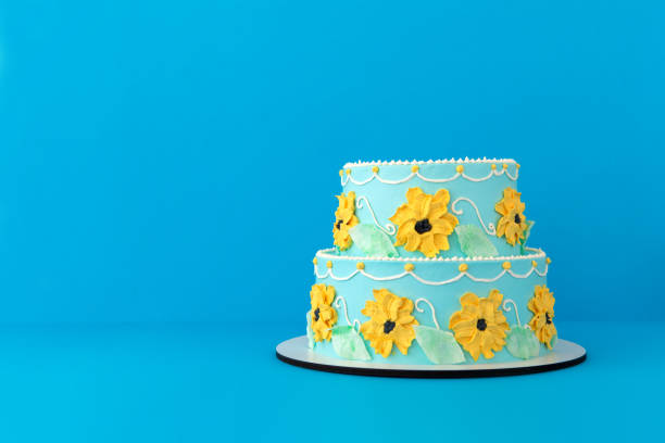 Colorful tiered festive birthday cake decorated with yellow flowers and green leaves made of cream on bright blue background