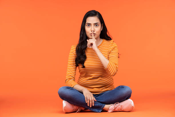 retrato de una mujer joven sentada aislada sobre fondo naranja - finger on lips whispering secrecy women fotografías e imágenes de stock