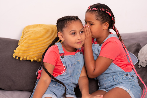 Little girl whispering in the sister's ear. Daughters wearing the same overalls and t-shirt, sitting on the sofa in the living room.