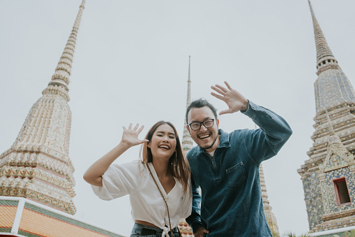 Southeast asian joyful young adult couple tourist looking at camera for taking photo, waving hand for greeting and smiling in front of Stupa of Wat Pho, The famous and ancient temple in Thailand on vacation