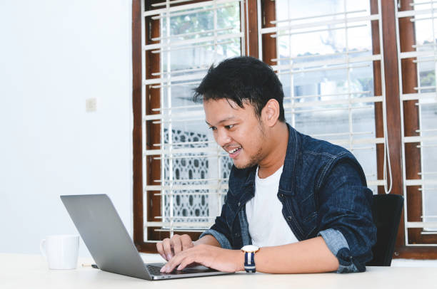 istock Young Asian man feeling happy and smile when work laptop on table. Indonesian man wearing blue shirt. 1330854886