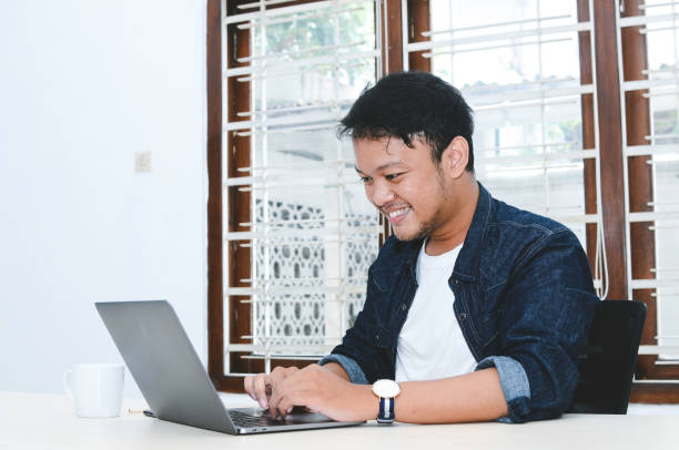 istock Young Asian man feeling happy and smile when work laptop on table. Indonesian man wearing blue shirt. 1330854906