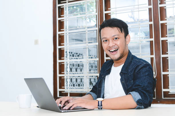 istock Young Asian man feeling happy and smile when work laptop on table. Indonesian man wearing blue shirt. 1330854977