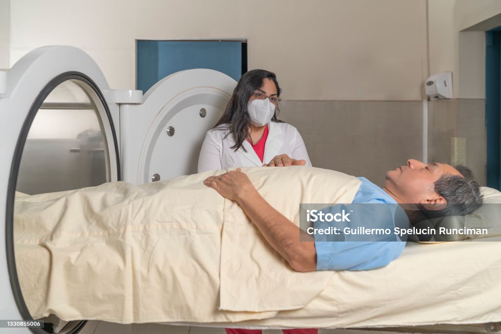 Doctor with mask talking to a patient lying Doctor with mask talking to a patient lying on a stretcher in front of a hyperbaric treatment machine. Hyperbaric Medicine Stock Photo