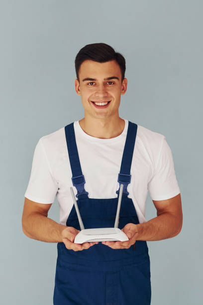 istock Holds wifi router. Male worker in blue uniform standing inside of studio against white background 1330859134
