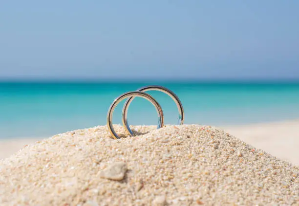 Pair of white gold wedding ring bands jewelry in sand on tropical desert island beach during summer with blue ocean background