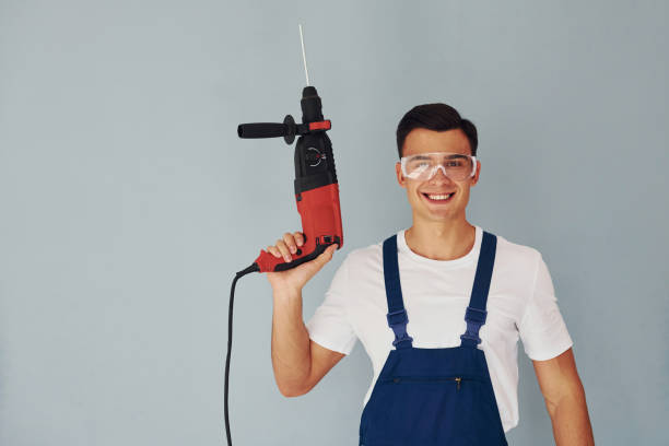 istock In protective eyewear and with drill in hands. Male worker in blue uniform standing inside of studio against white background 1330863613